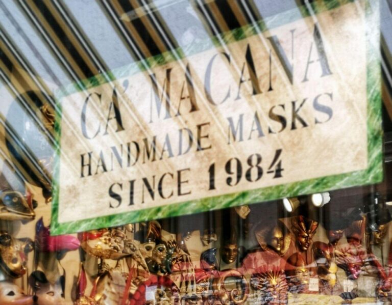 Shopfront window of Ca Macana in Venice showing the Venetian Carnival masks and the buildings and canals reflected in the window