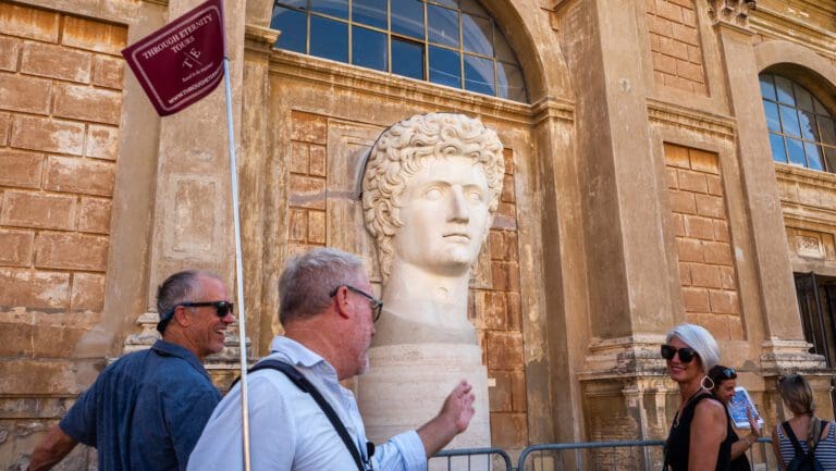 Tourists walk past a Roman emperor statue outside a historic building.