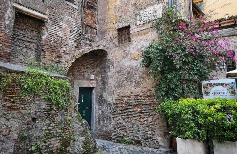 Quiet corner of the Jewish Ghetto in Rome showing an anceint brick building with a green wooden door and the sign to a local restaurant, all surrounded by foliage.